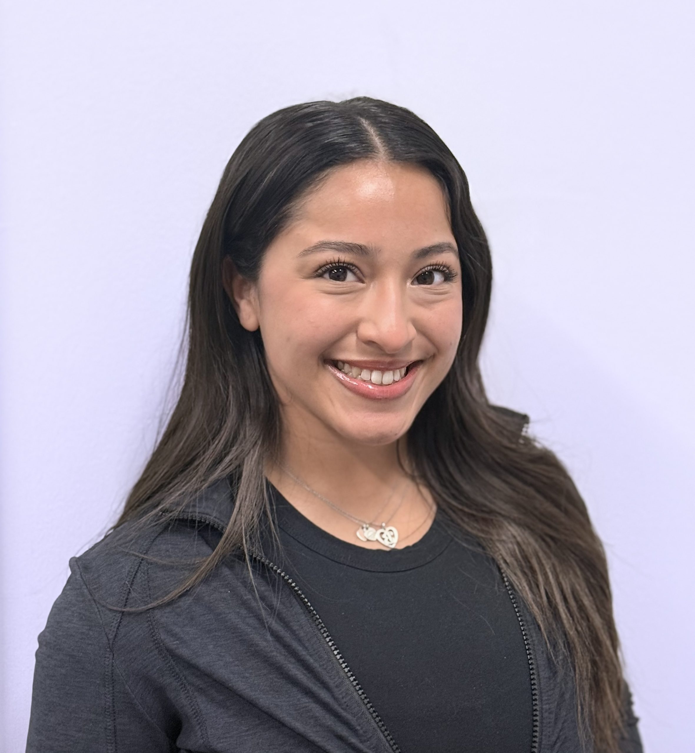 A woman with long dark hair smiles confidently at the camera, exuding a professional aura in a black top and jacket, against a plain light background—a true leader ready to support her administrative team.